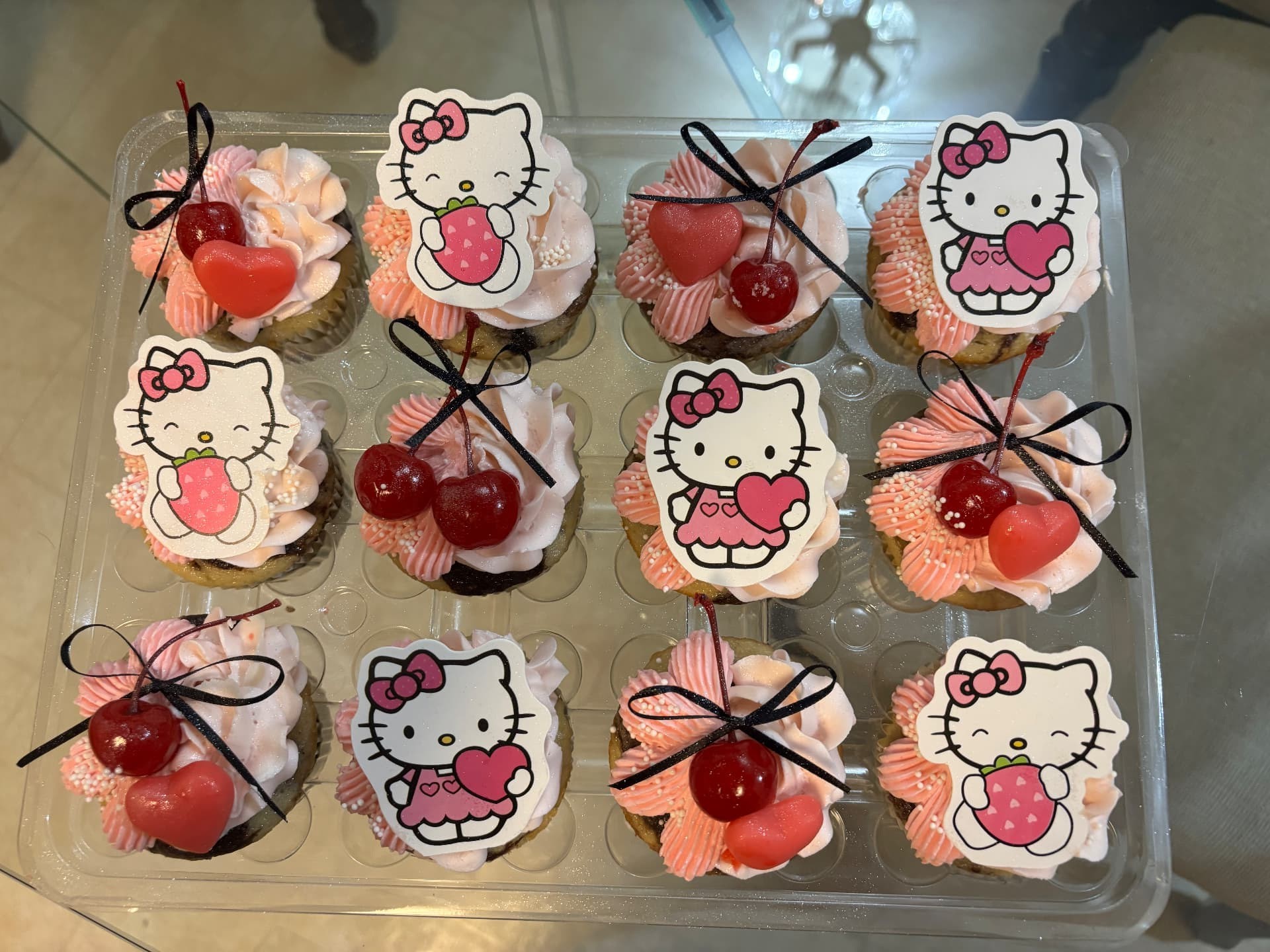 A real BakeToGive dozen cupcakes with pink and white buttercream swirls, pearl sprinkles, cherries with black bows and character toppers